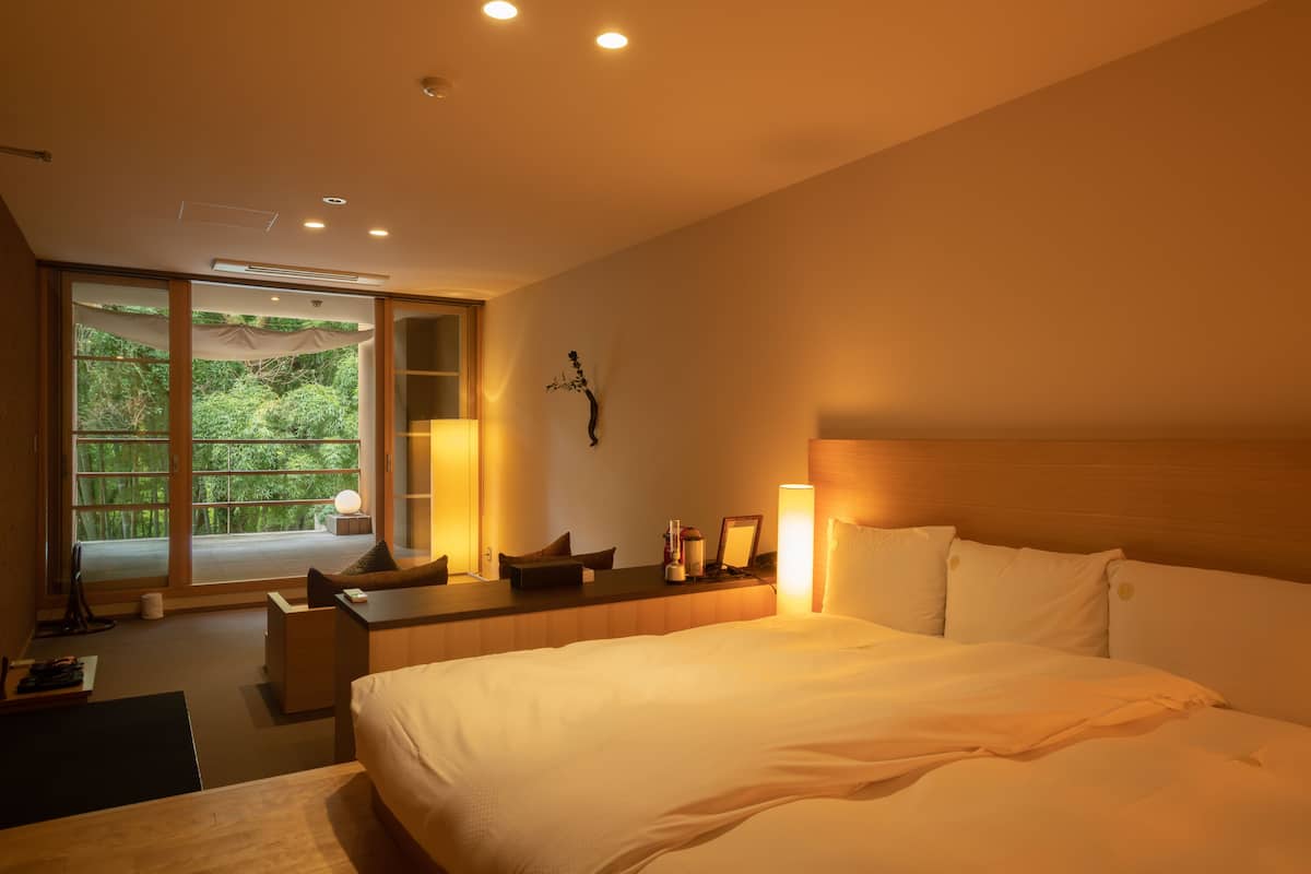 A modern hotel room with a large bed, a desk, and a view of greenery through the window.