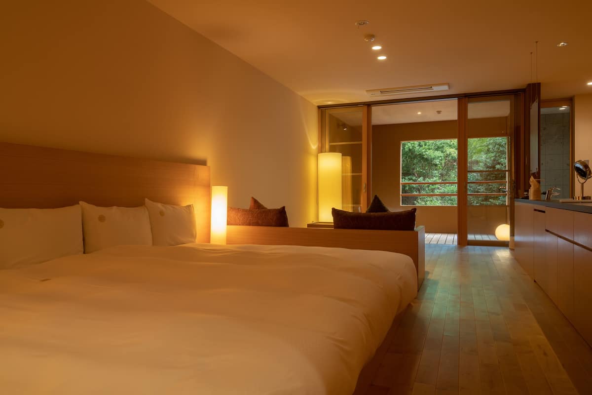 A modern hotel room with a large bed, a wooden headboard, a bedside table with a lamp, and a sliding glass door leading to a balcony with a view of greenery.