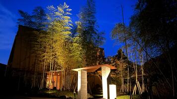 A rustic wooden structure with a sloped roof, surrounded by tall trees and a building in the background.