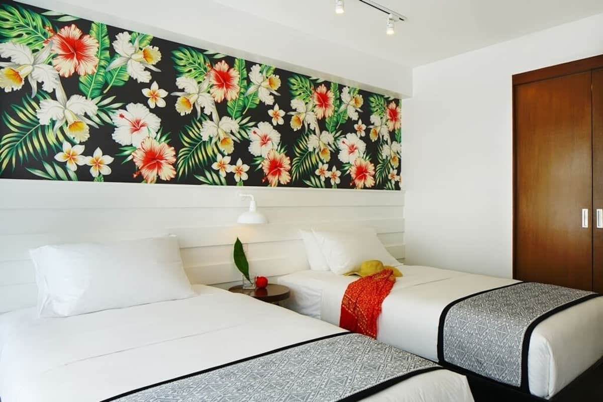 A hotel room with two beds, a large floral wall mural, a wooden wardrobe, and a nightstand with a lamp.