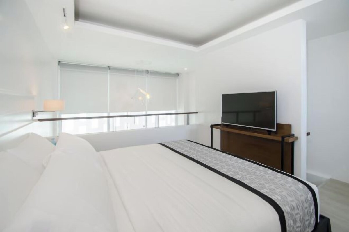 Una habitación de hotel moderna con una cama grande, un televisor de pantalla plana sobre un soporte de madera y un ventanal con vistas a la ciudad.