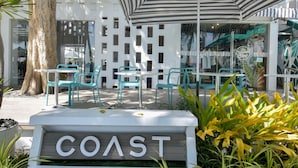 Un espacio al aire libre con temática costera, que incluye una señalización que dice "COAST", sillas turquesa y un paraguas a rayas.