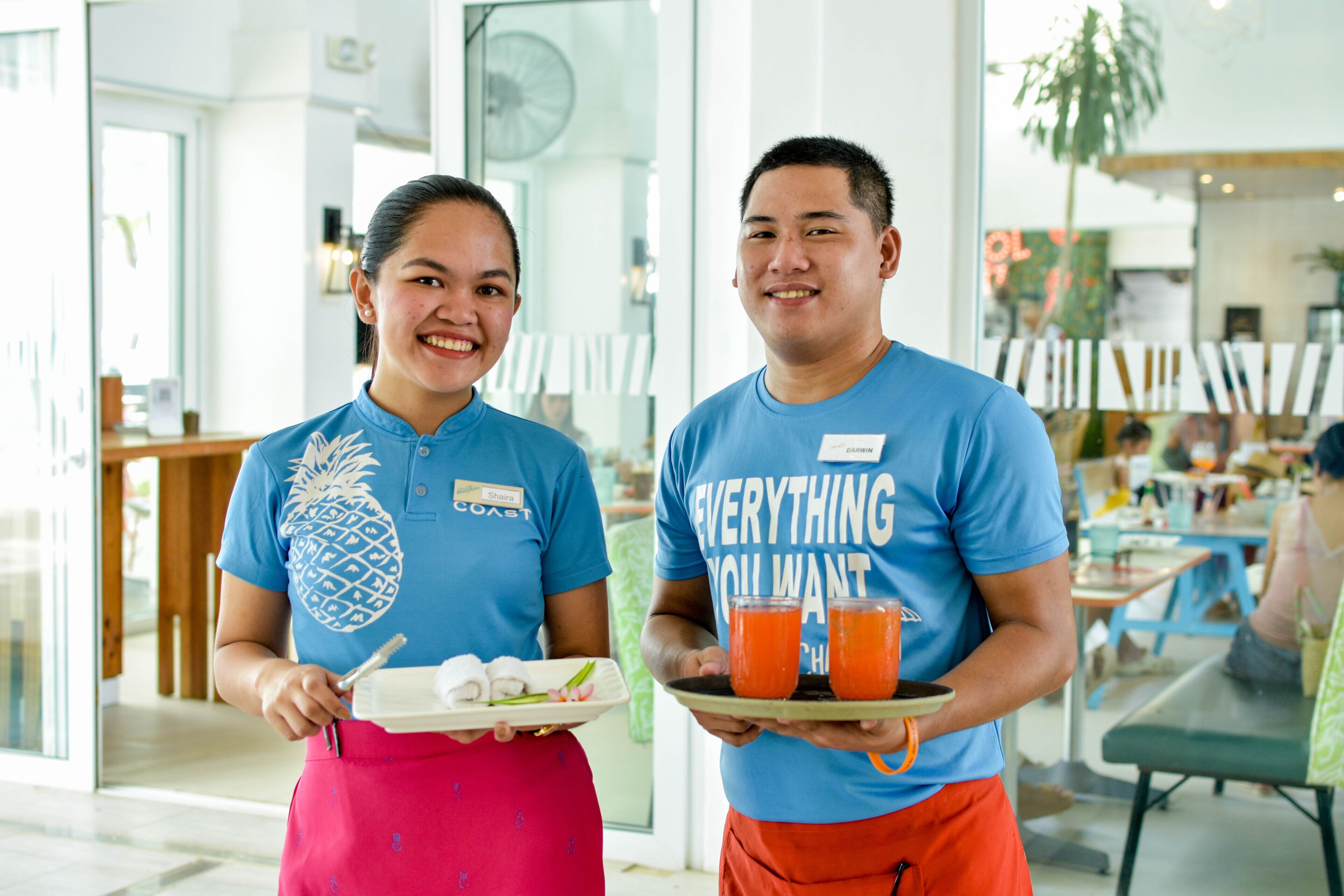 Two staff members in a bright indoor setting, one holding a tray with food and drinks, and the other holding a tray with a pineapple design.