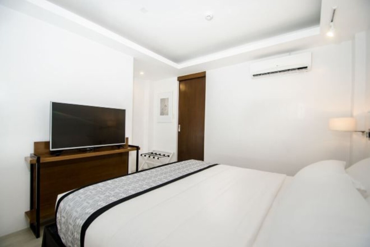 A hotel room with a large bed, a television on a wooden stand, a white chair, and a door.