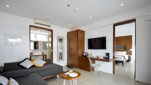 Una habitación de hotel moderna con un sofá gris, una pequeña mesa redonda, un televisor, un escritorio y una cama al fondo.