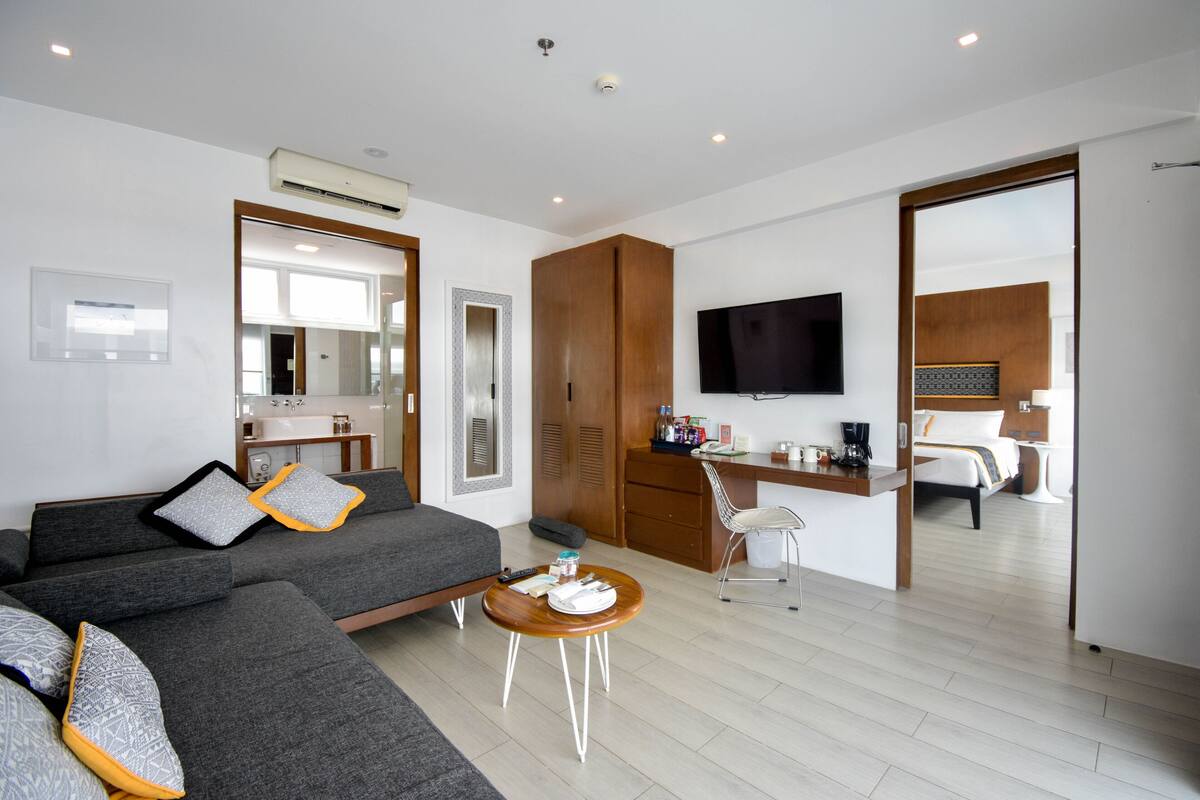 A modern hotel room with a grey sofa, a small round table, a TV, a desk, and a bed in the background.