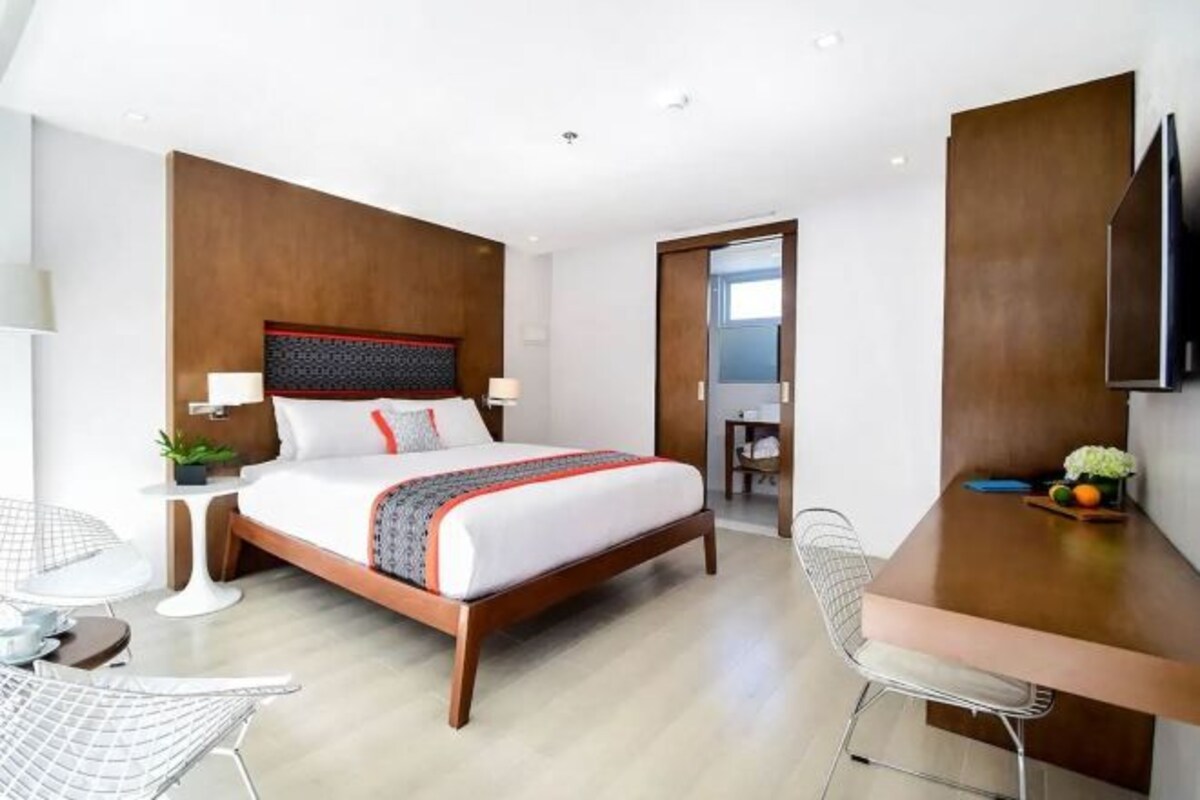A modern hotel room with a wooden headboard, a large bed, a desk, and a chair.