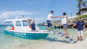 Un bote llamado 'COAST' atracado en una playa con agua cristalina, una palmera y una estructura de techo de paja al fondo.