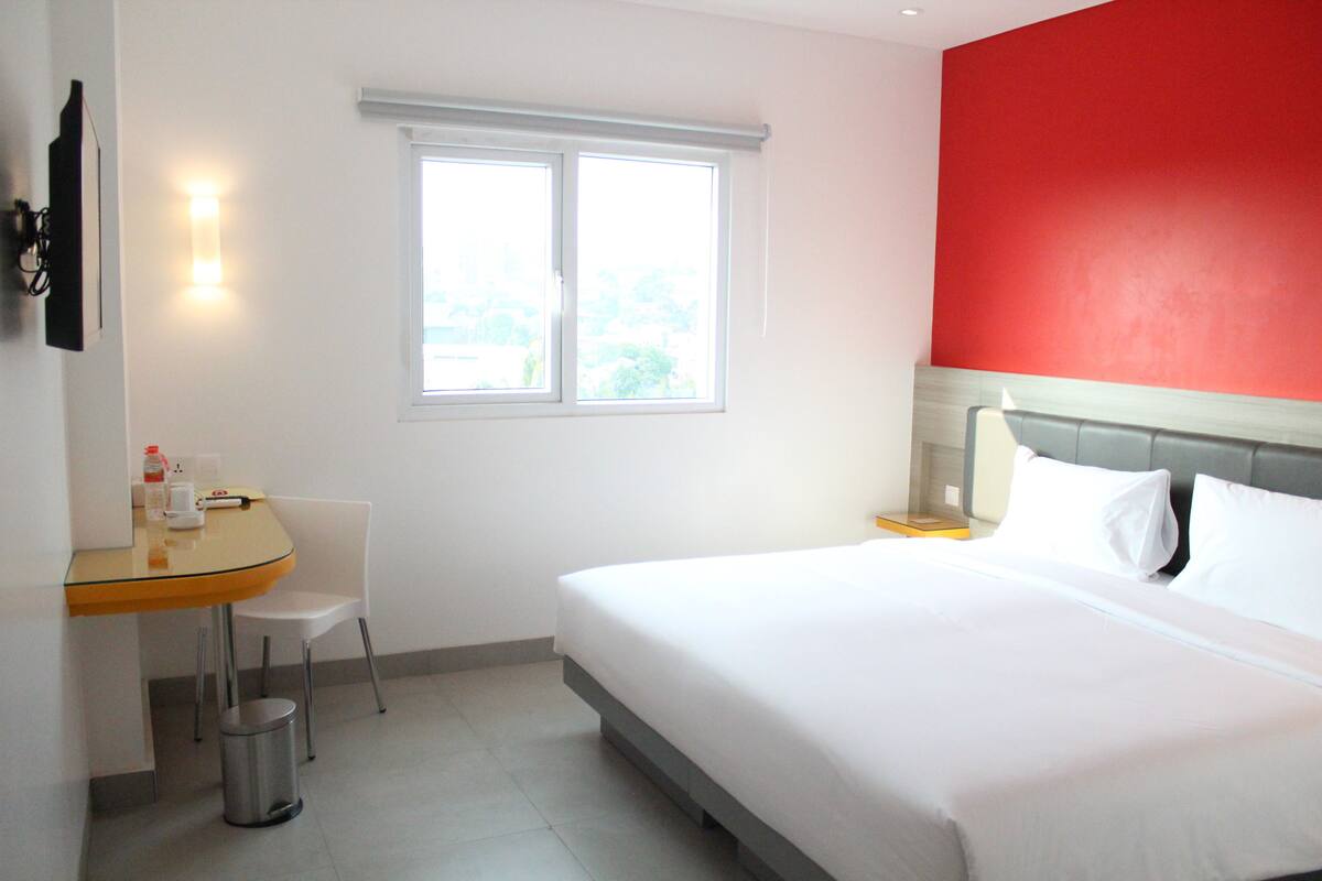 A hotel room with a bed, a desk with a chair, a window, and a red accent wall.