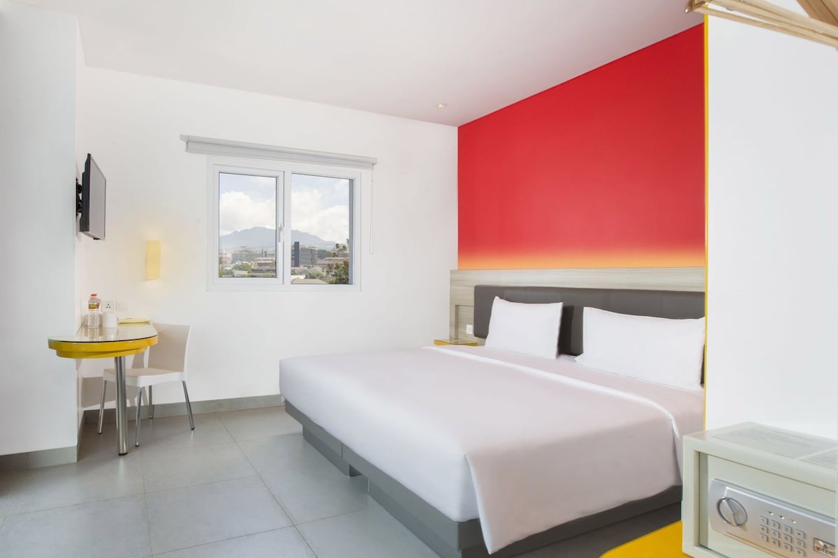 A hotel room with a bed, a desk, a chair, a window with a city view, and a red accent wall.
