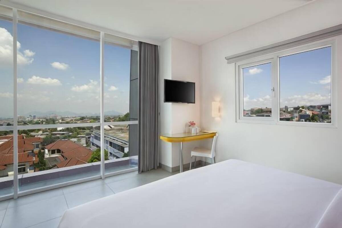 A hotel room with a bed, a desk, a TV, and large windows offering a city view.