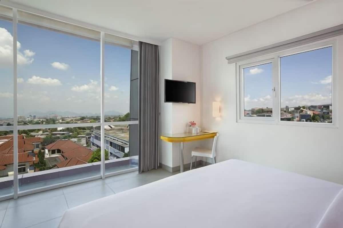 A hotel room with a bed, a desk, a TV, and large windows offering a city view.