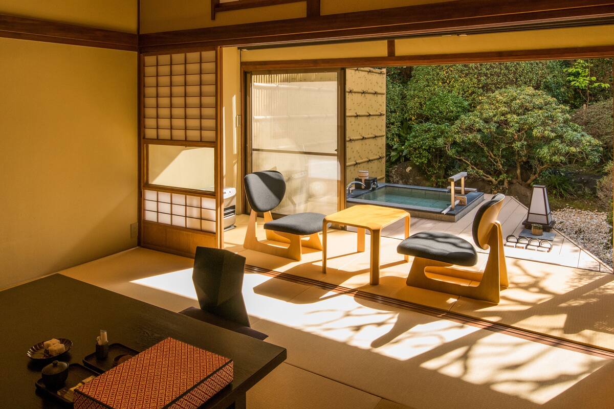 A traditional Japanese room with tatami flooring, a low table, two chairs, and a view of a garden and outdoor hot tub.