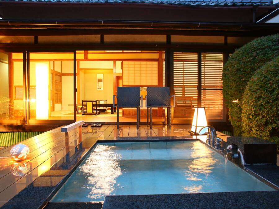 A swimming pool with a wooden deck, a lounge area with chairs, and a view of a traditional Japanese building.