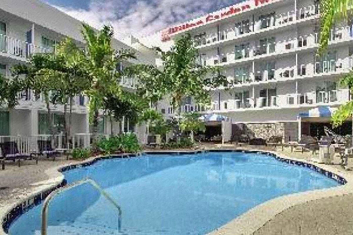 Zona de piscina hotelera con una piscina azul cristalina, rodeada de tumbonas y sombrillas, y un edificio con balcones al fondo.