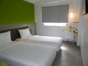 Habitación de hotel con dos camas, un escritorio pequeño, una ventana con persiana y una pared de acento verde.