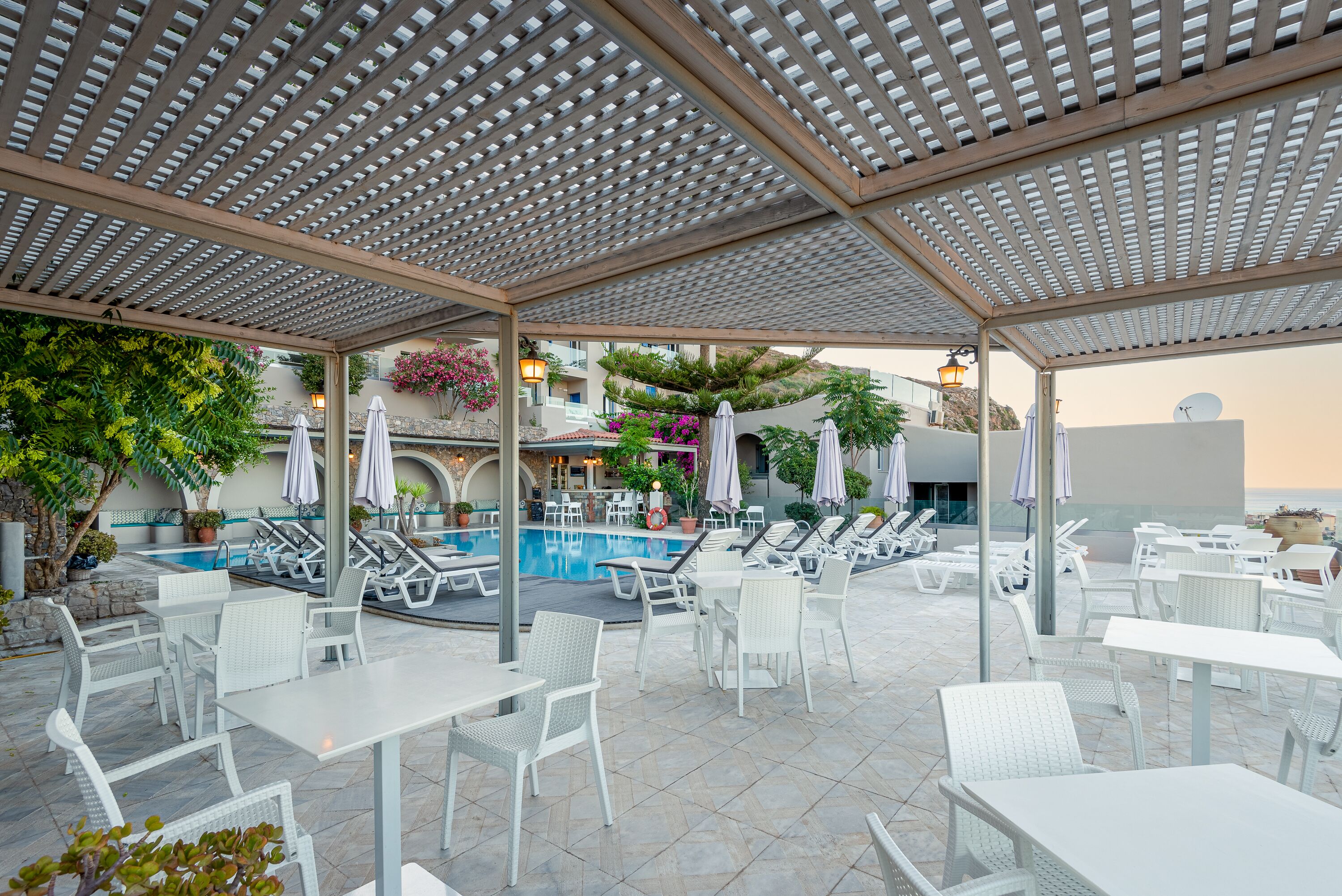 An outdoor pool area with white lounge chairs and tables, a covered patio with a pergola, and a view of the ocean.