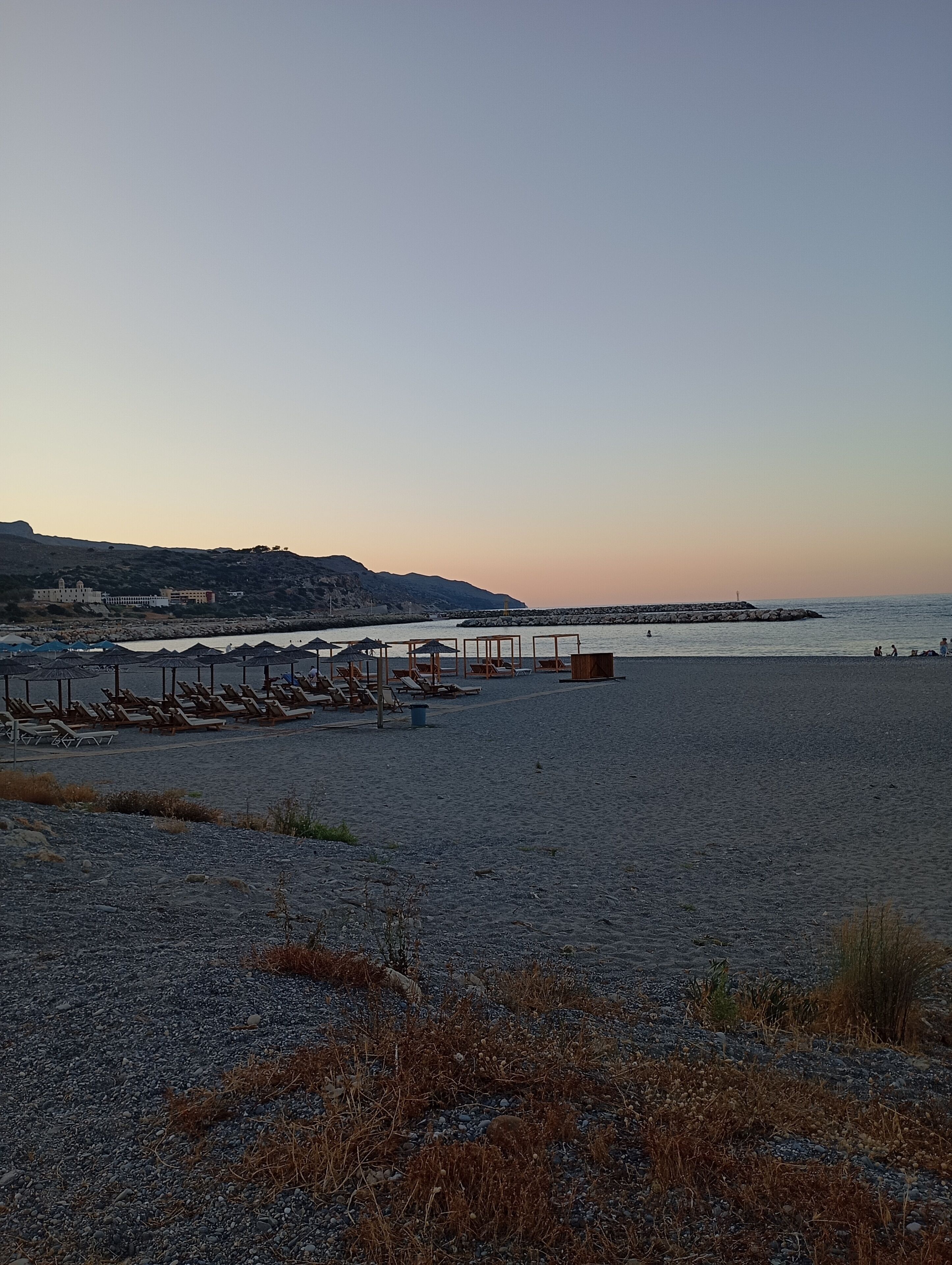 Una playa con tumbonas y sombrillas, un muelle que se adentra en el agua, y un edificio al fondo.