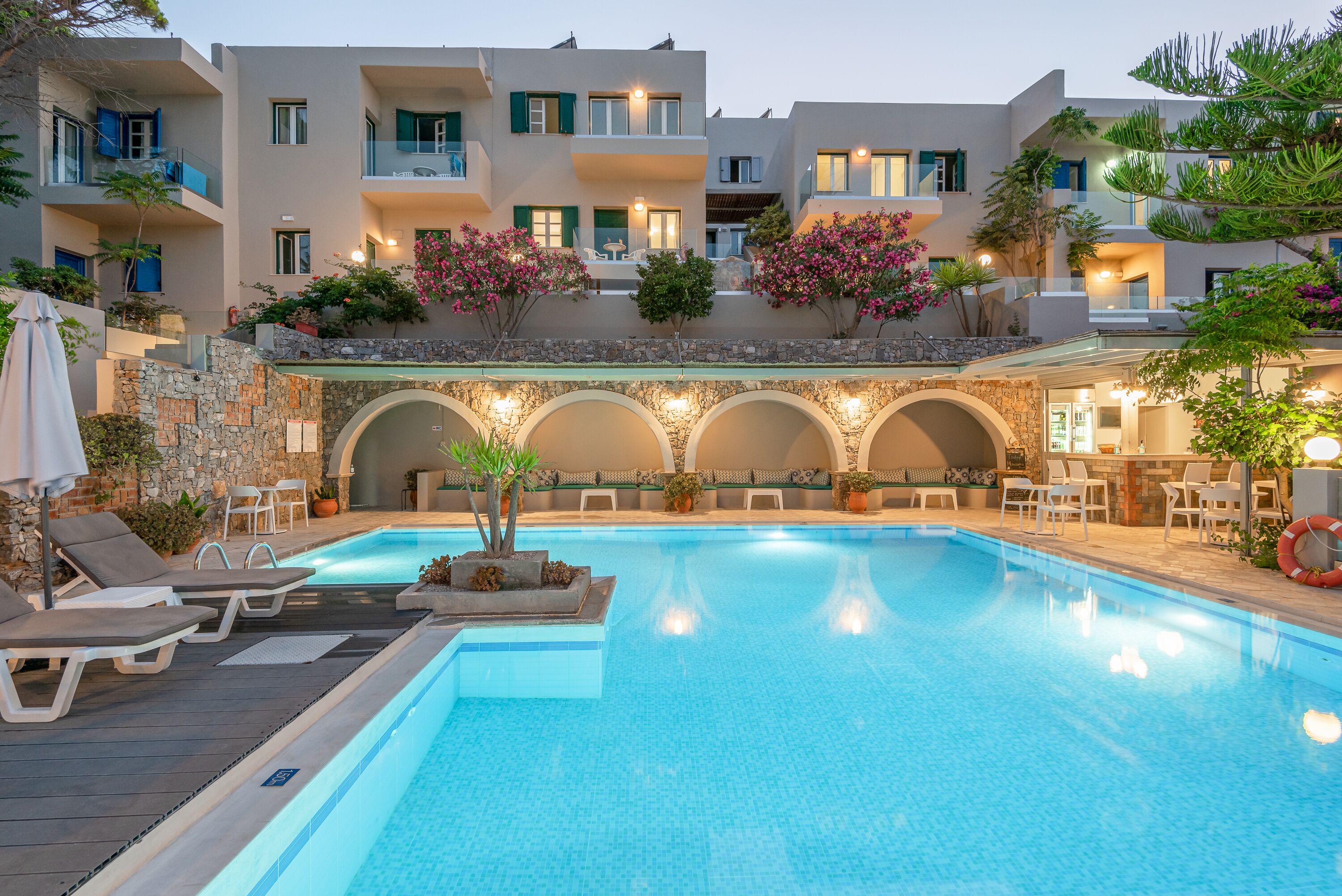 Una piscina con sillas de descanso, un muro de piedra y un edificio de varios pisos con balcones y plantas en macetas.