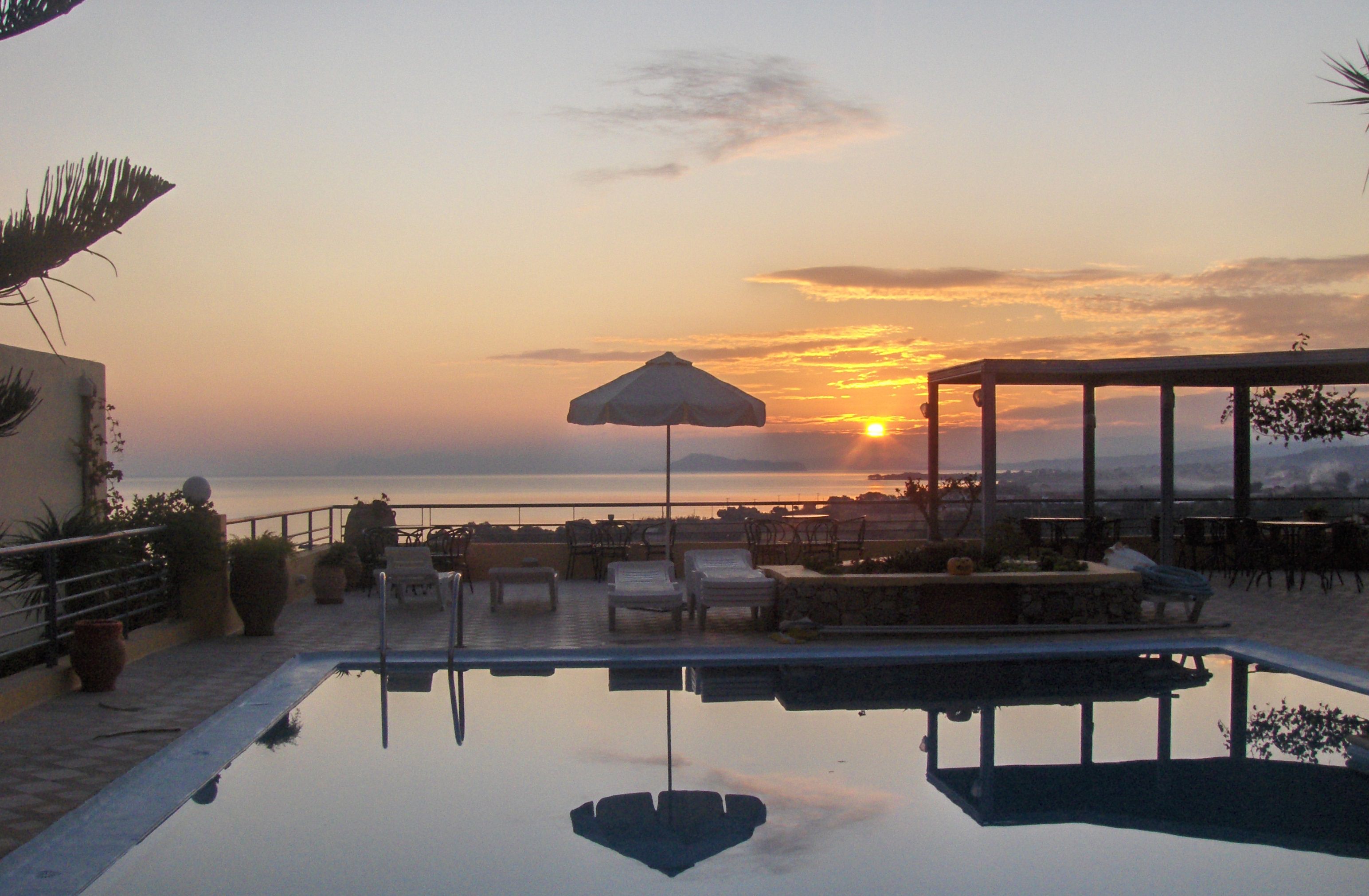 Zona junto a la piscina con sillones y una sombrilla, con vista a la puesta de sol sobre el mar.