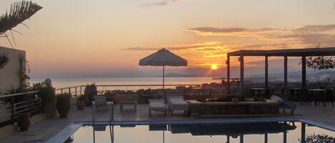 Zona junto a la piscina con sillones y una sombrilla, con vista a la puesta de sol sobre el mar.