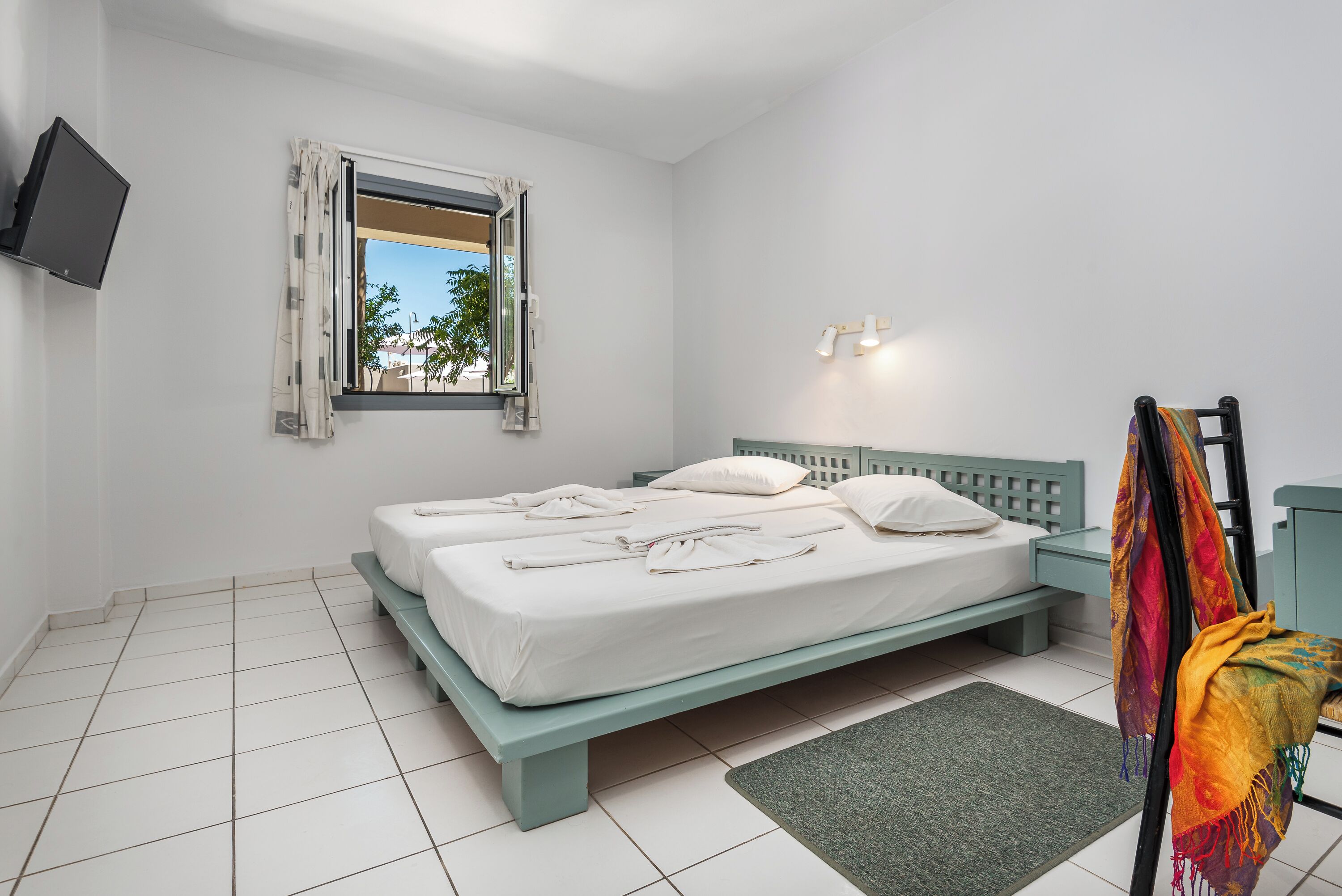 Quarto com cama, televisão fixada na parede, janela com cortinas e mesa de cabeceira com abajur.