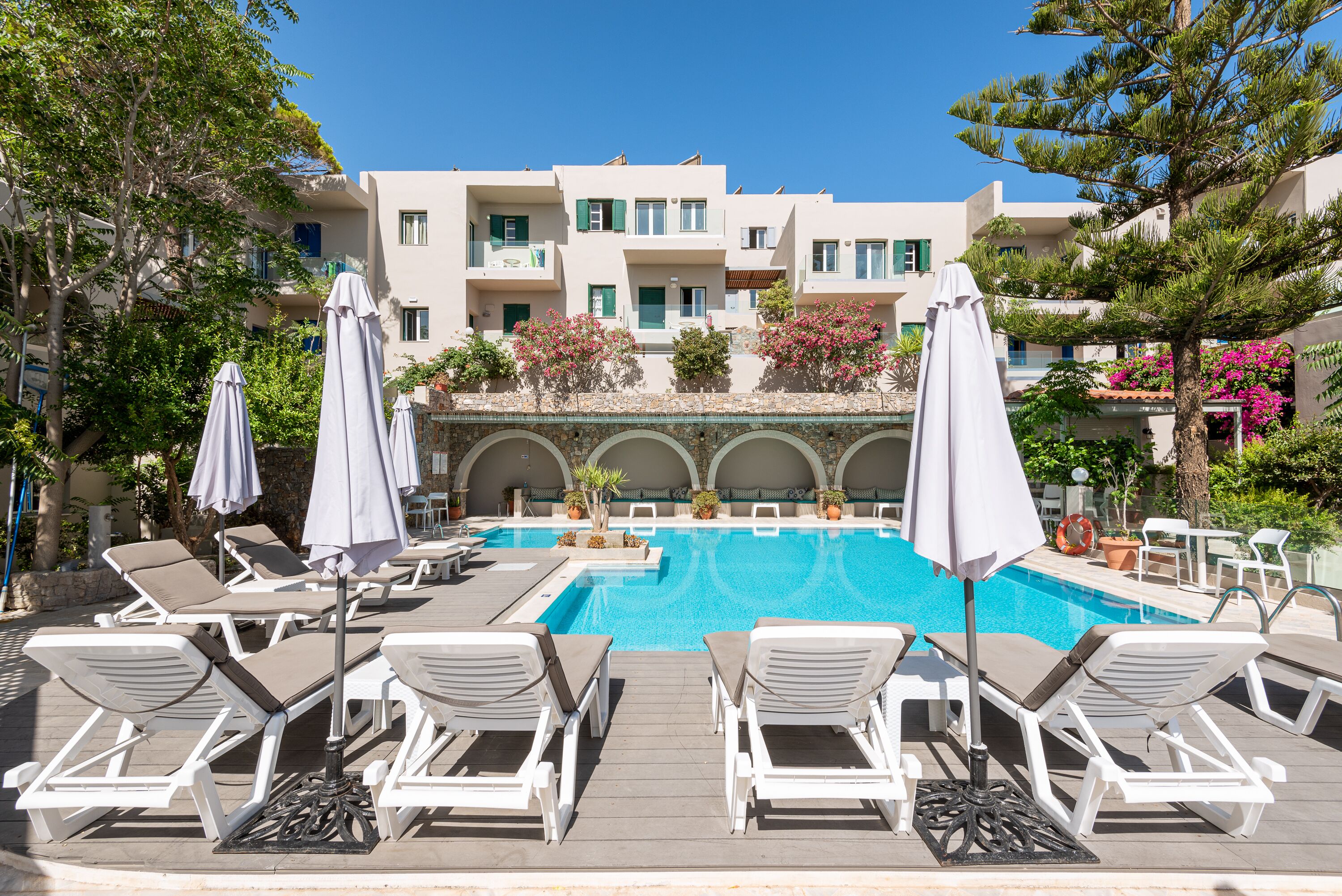 Un área junto a la piscina con sillones de descanso y sombrillas, una piscina azul cristalina y un edificio con balcones al fondo.