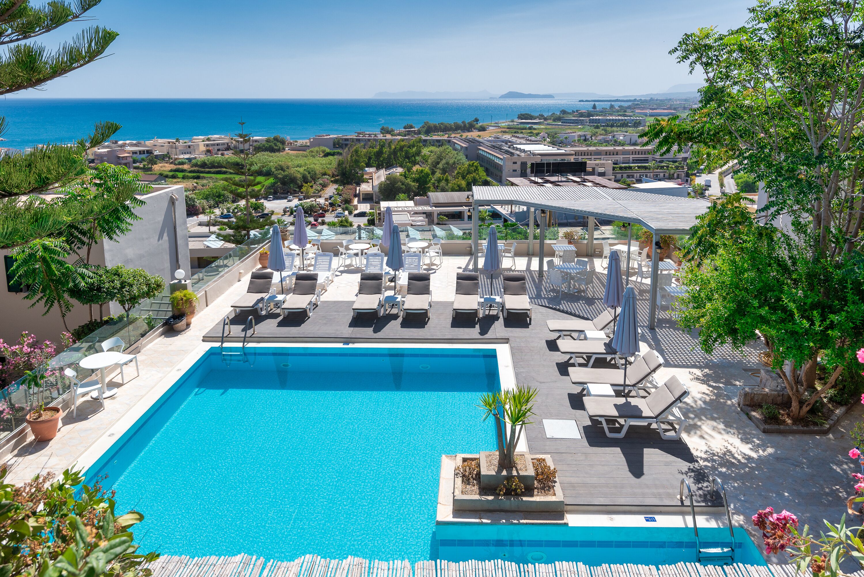 A rooftop pool with lounge chairs and umbrellas, surrounded by greenery and overlooking a coastal cityscape.