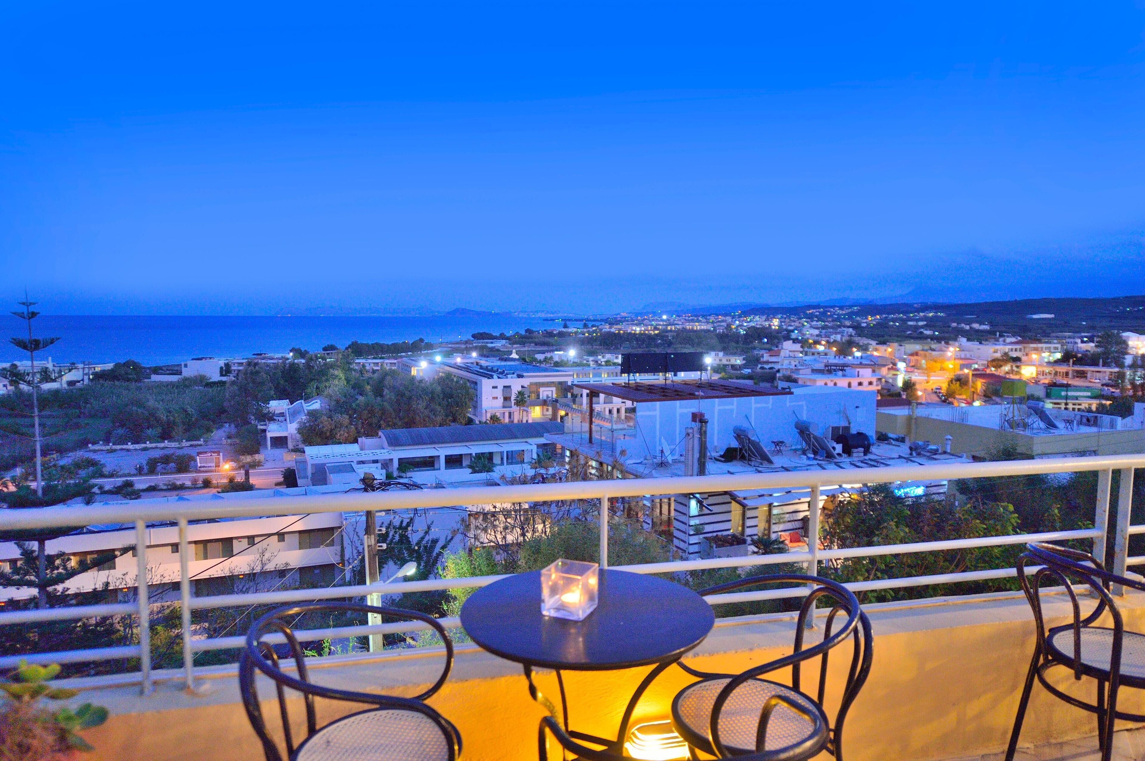 A balcony with a table and chairs, overlooking a coastal cityscape at dusk.