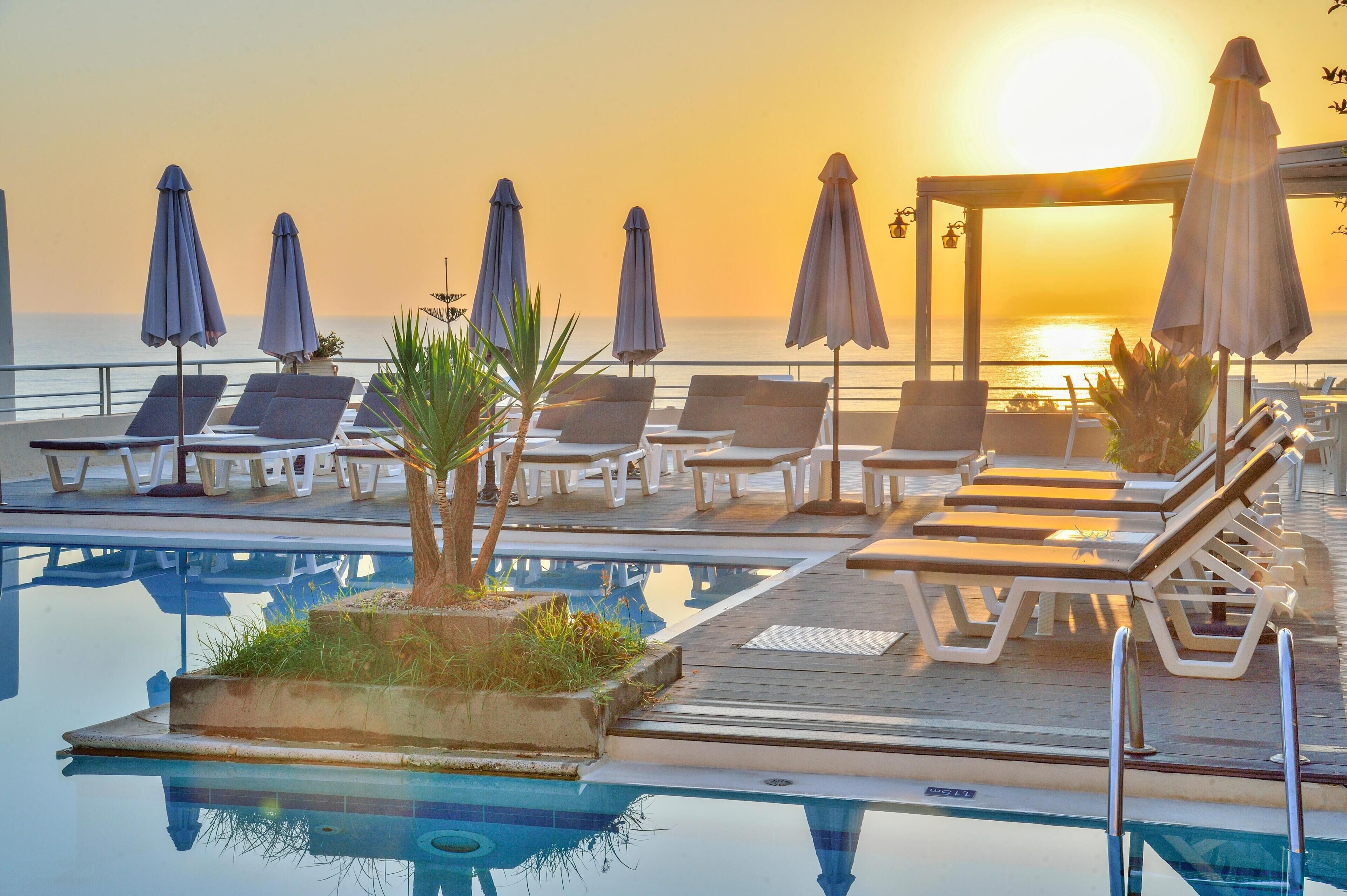 Zona junto a la piscina con sillones de descanso y sombrillas, una terraza de madera y vista al océano al atardecer.