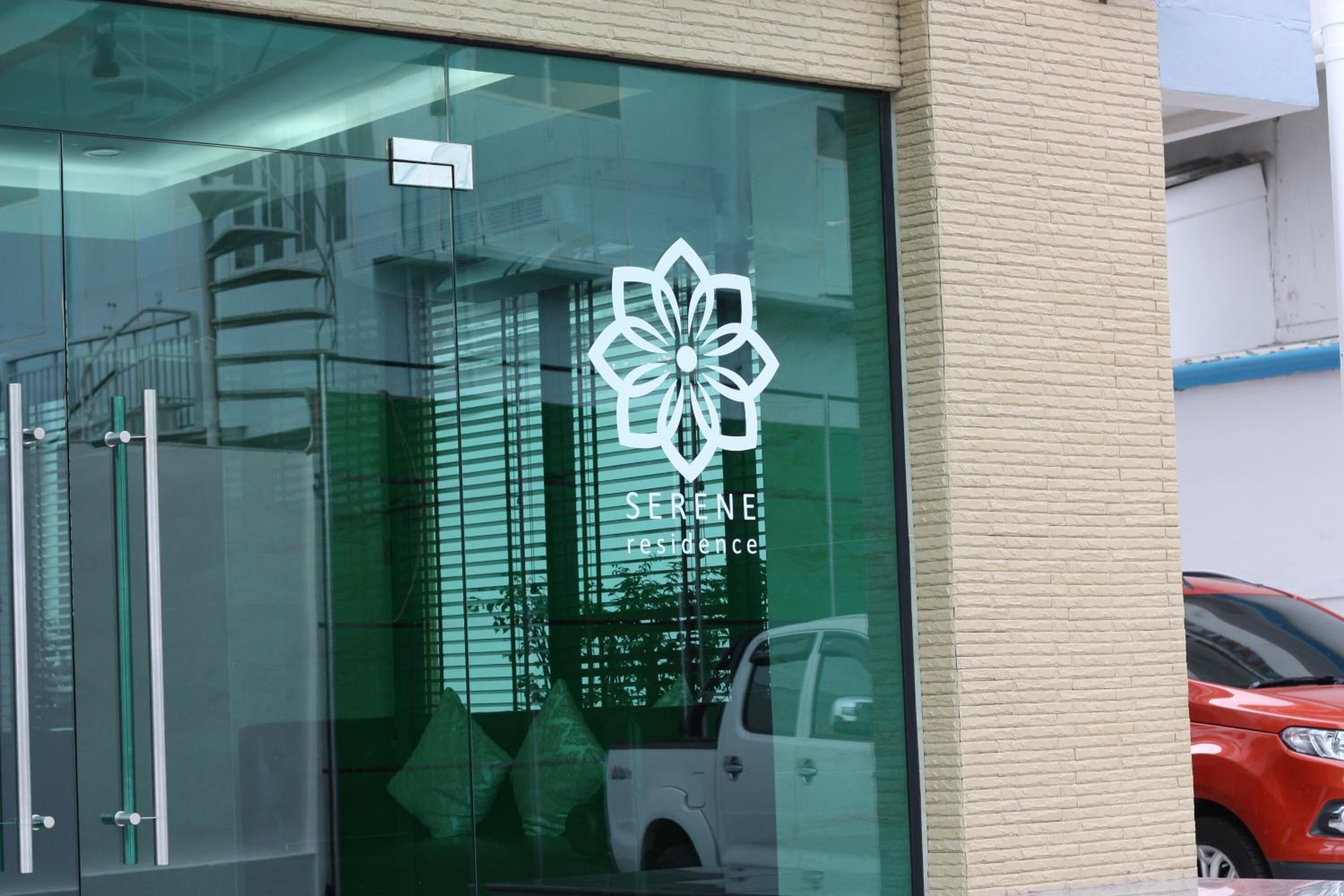 Un edificio con fachada de vidrio, un logotipo con el texto "SERENE residence", y el reflejo de un coche y un edificio en el cristal.