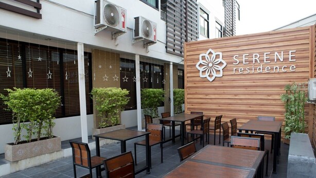 An outdoor seating area with tables and chairs, a wooden wall with the text "SERENE residence," and air conditioning units mounted on the wall. - Serene Residence (Kanchanaburi)