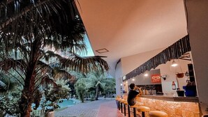 Un área de bar interior con mostrador de piedra y taburetes, rodeada de plantas tropicales y un camino de ladrillo.