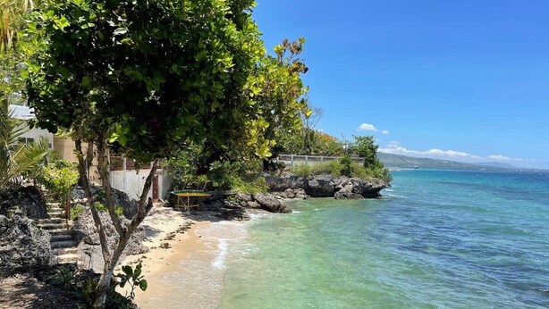 Una playa con agua cristalina, una orilla arenosa y un promontorio rocoso con una silla y una mesa.