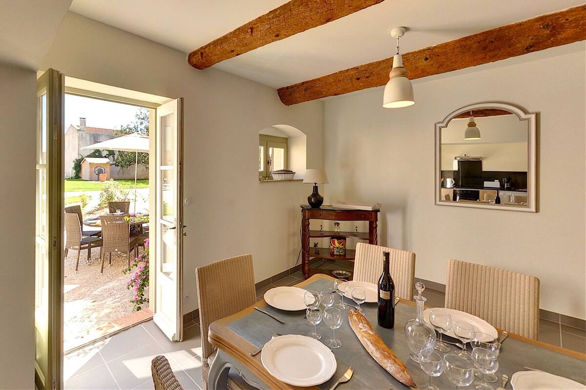 Un coin repas avec une table dressée pour quatre personnes, une bouteille de vin et une vue sur le jardin par une porte ouverte.