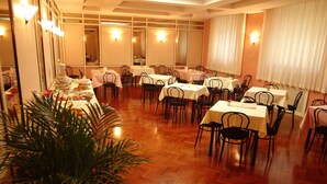 Uma sala de jantar com várias mesas e cadeiras, uma grande planta em vaso e luminárias na parede.