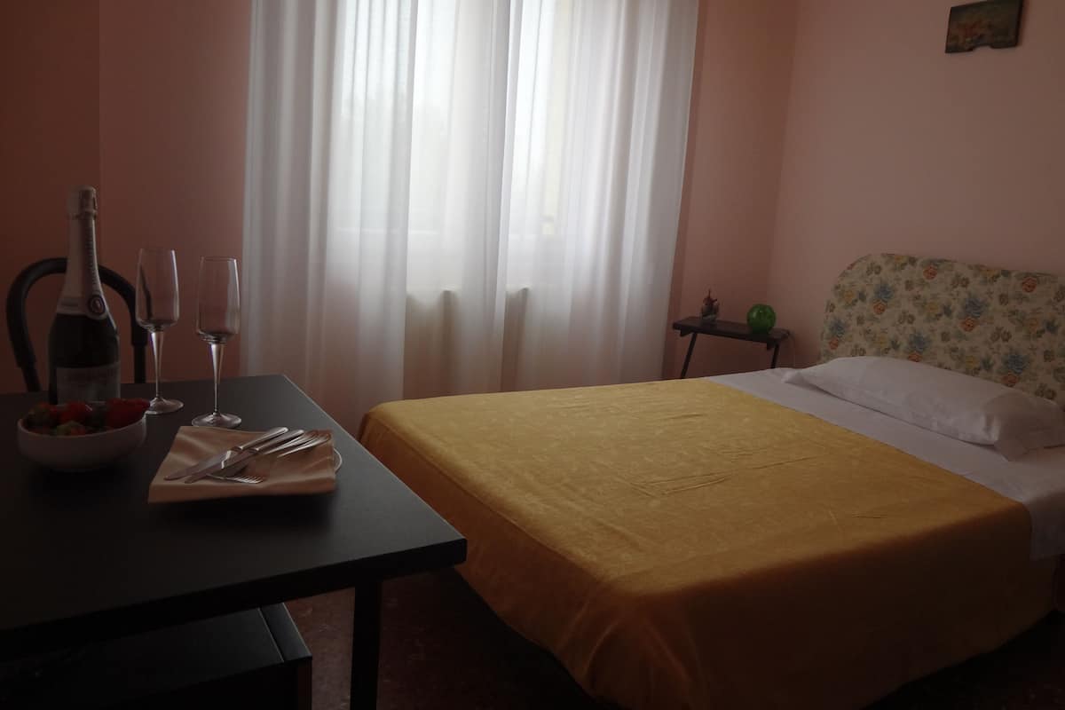 A hotel room with a bed, a table with a bottle and glasses, a bowl of fruit, and a small table with a vase and a green object.