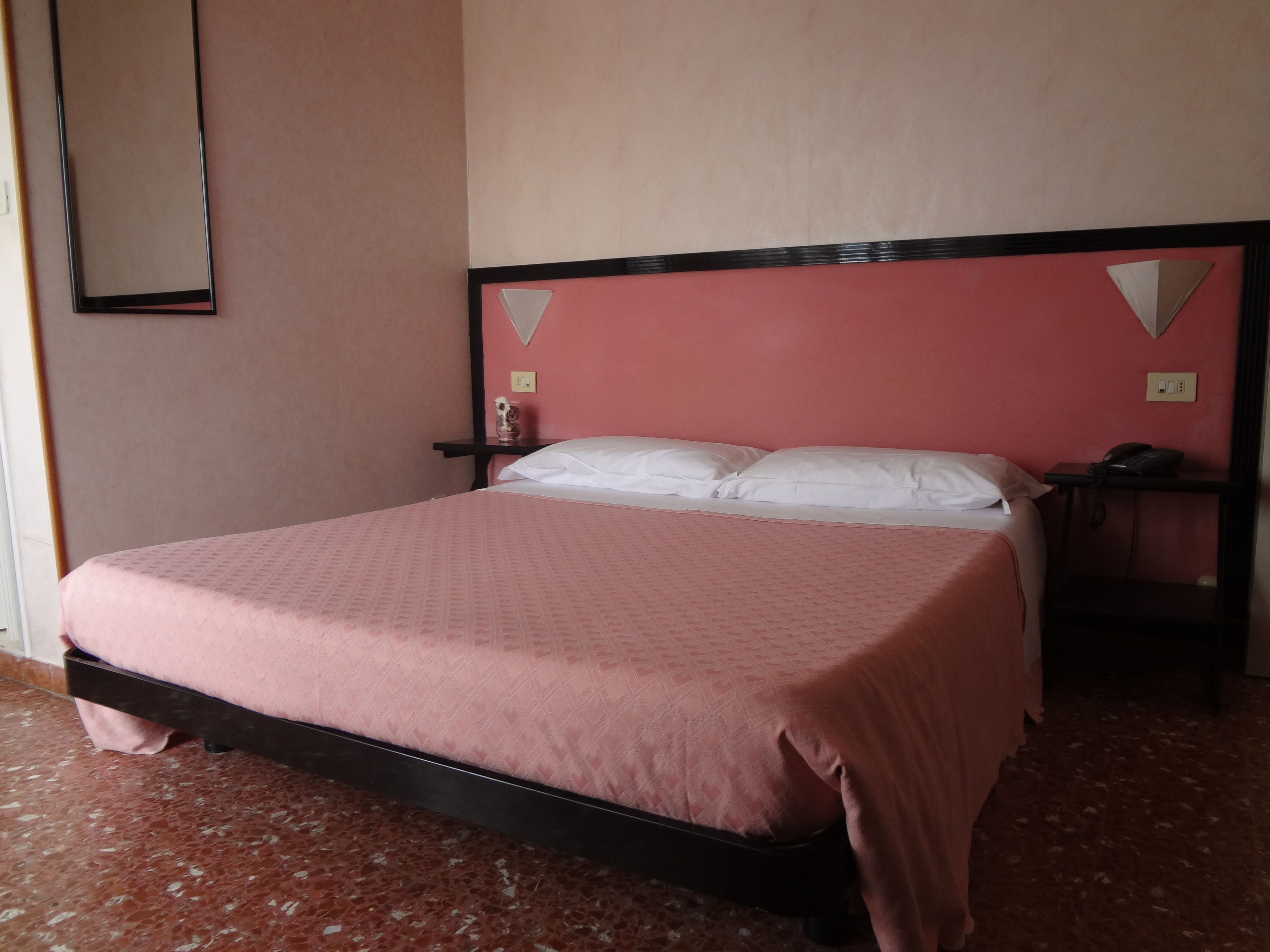 A bed with a pink bedspread, a wooden headboard, a nightstand with a phone, and a wall-mounted light switch.