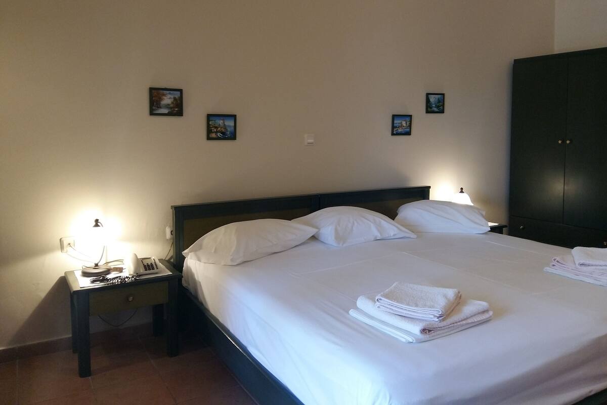 A neatly made bed with white linens, a bedside lamp, a small table with a phone and a lamp, and a dark-colored wardrobe.
