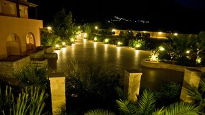 Vue nocturne d’une propriété située au bord de l’eau, avec des allées éclairées, des escaliers en pierre et une végétation luxuriante.