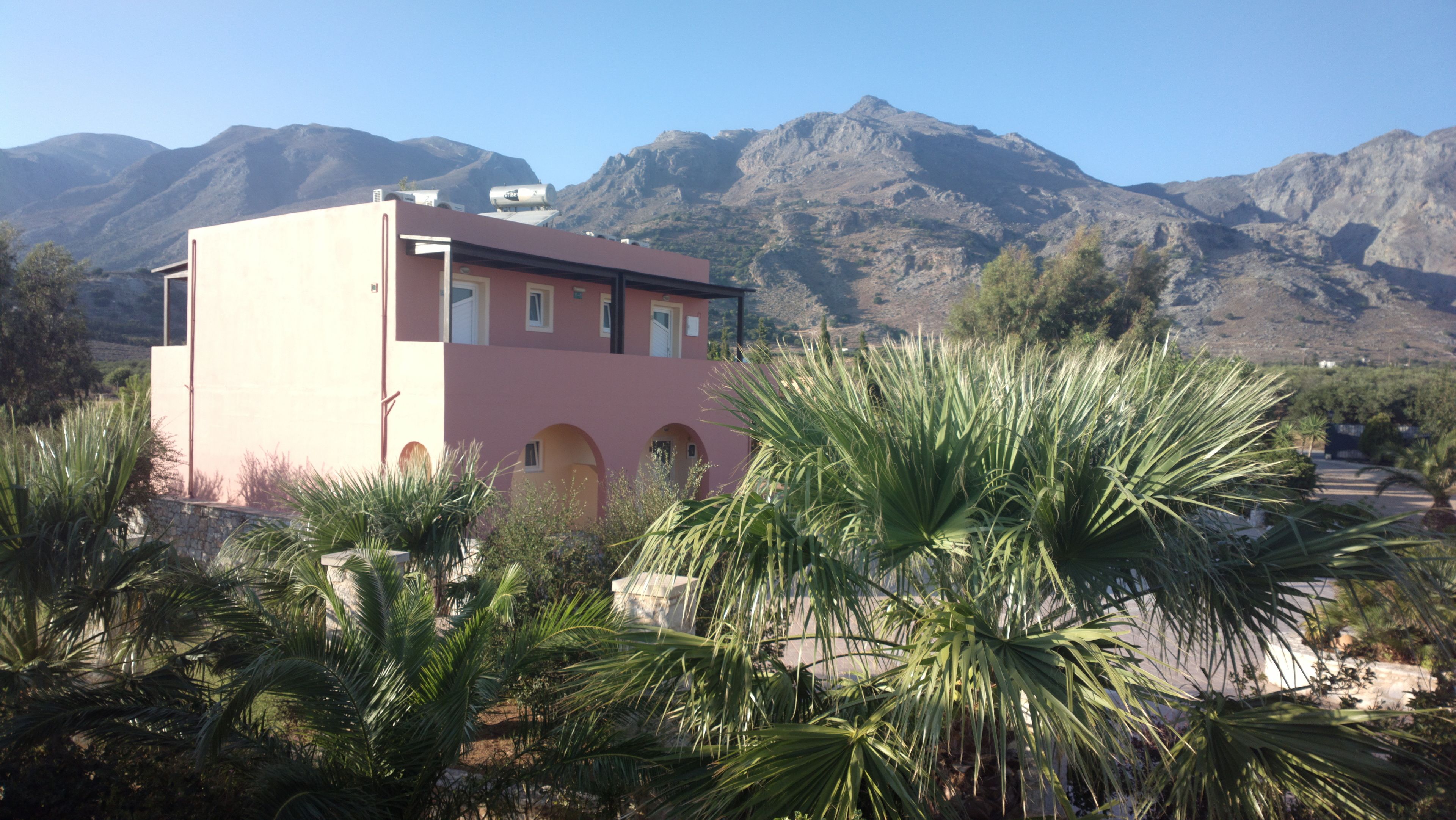 Un edificio rosa con ventanas arqueadas y techo plano, rodeado de palmeras y montañas al fondo.