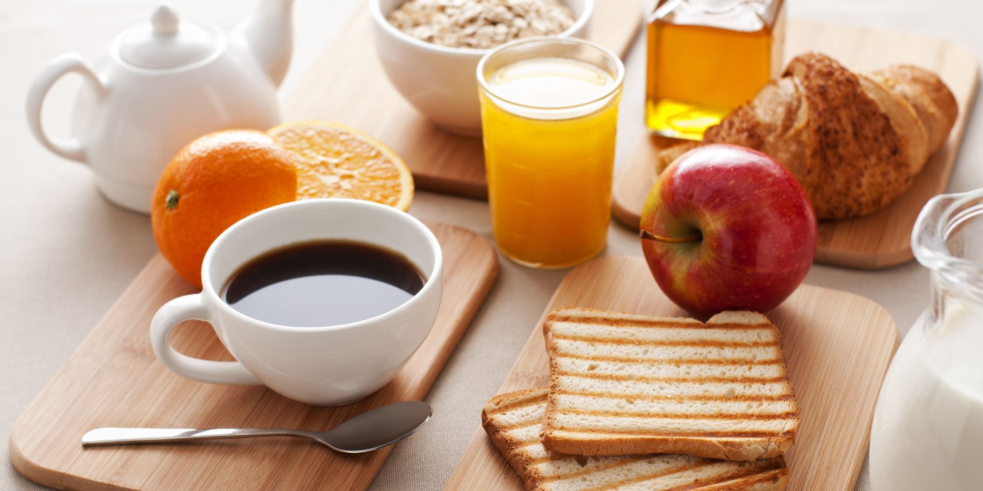 Een ontbijtpresentatie met een kop koffie, sinaasappelsap, toast, een appel, een croissant en een glas melk op een houten plank.