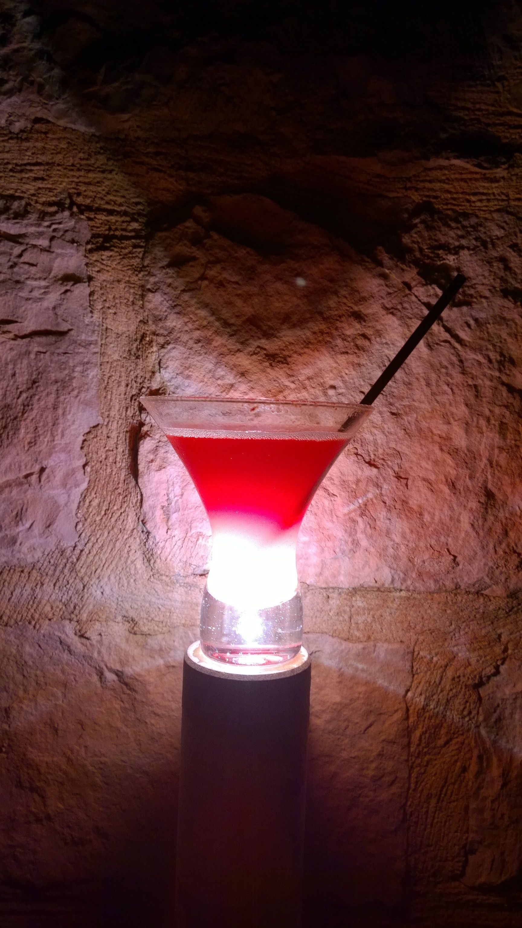 Una copa de martini con un líquido rojo sobre una base negra, frente a una pared de piedra con textura.