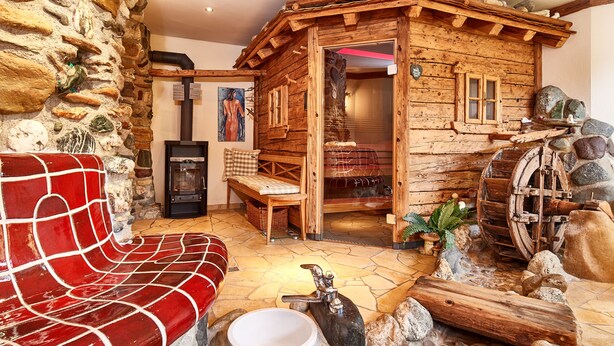 Interior rústico de una cabaña con pared de piedra, cabaña de madera, banco, chimenea y una noria decorativa.