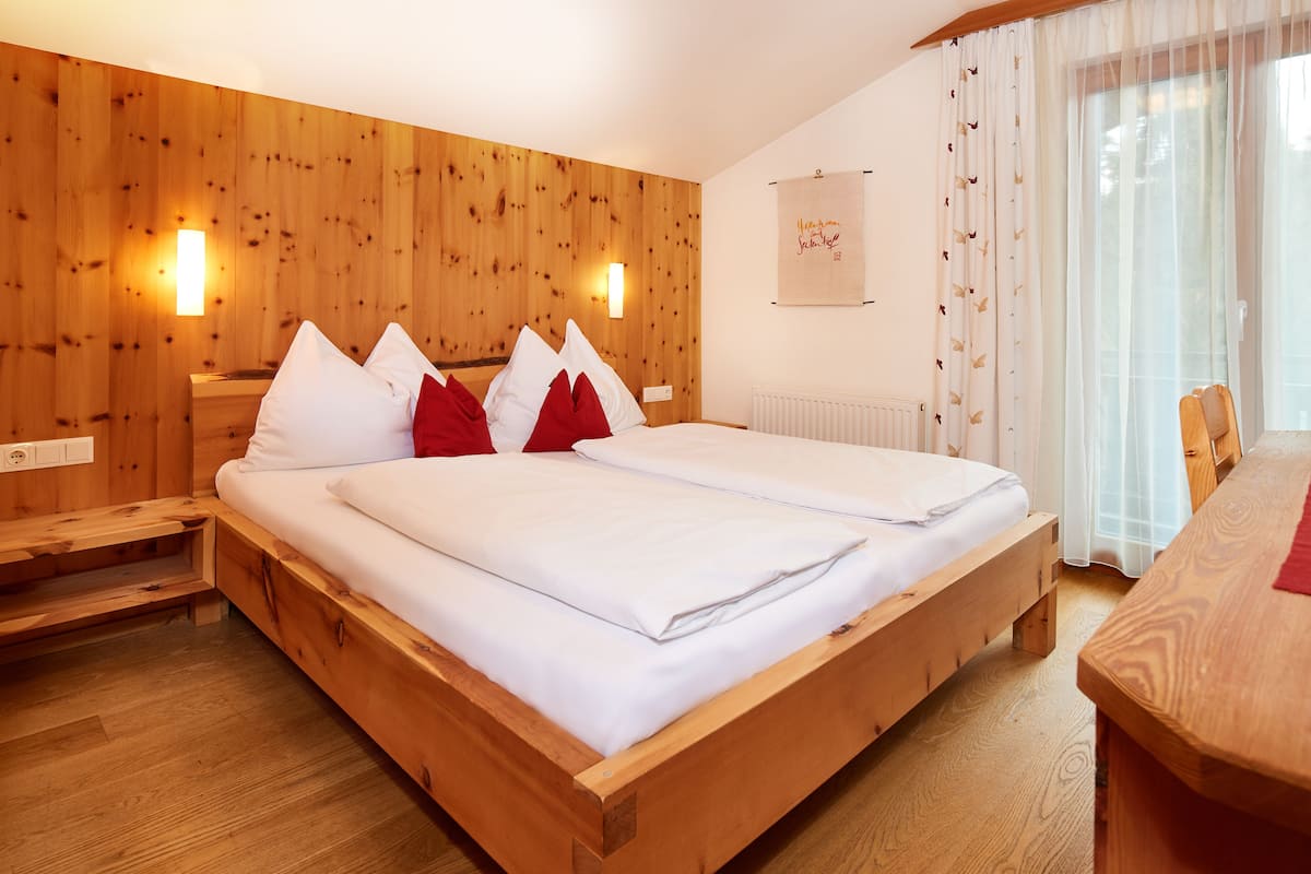 Un lit en bois avec des oreillers blancs et rouges, une table de chevet en bois, une table en bois et un luminaire fixé au mur.