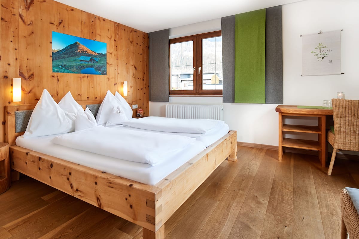 Un lit en bois avec du linge de lit blanc, une table de chevet en bois, une chaise en osier, une table, une fenêtre donnant sur des immeubles et un tableau accroché au mur.