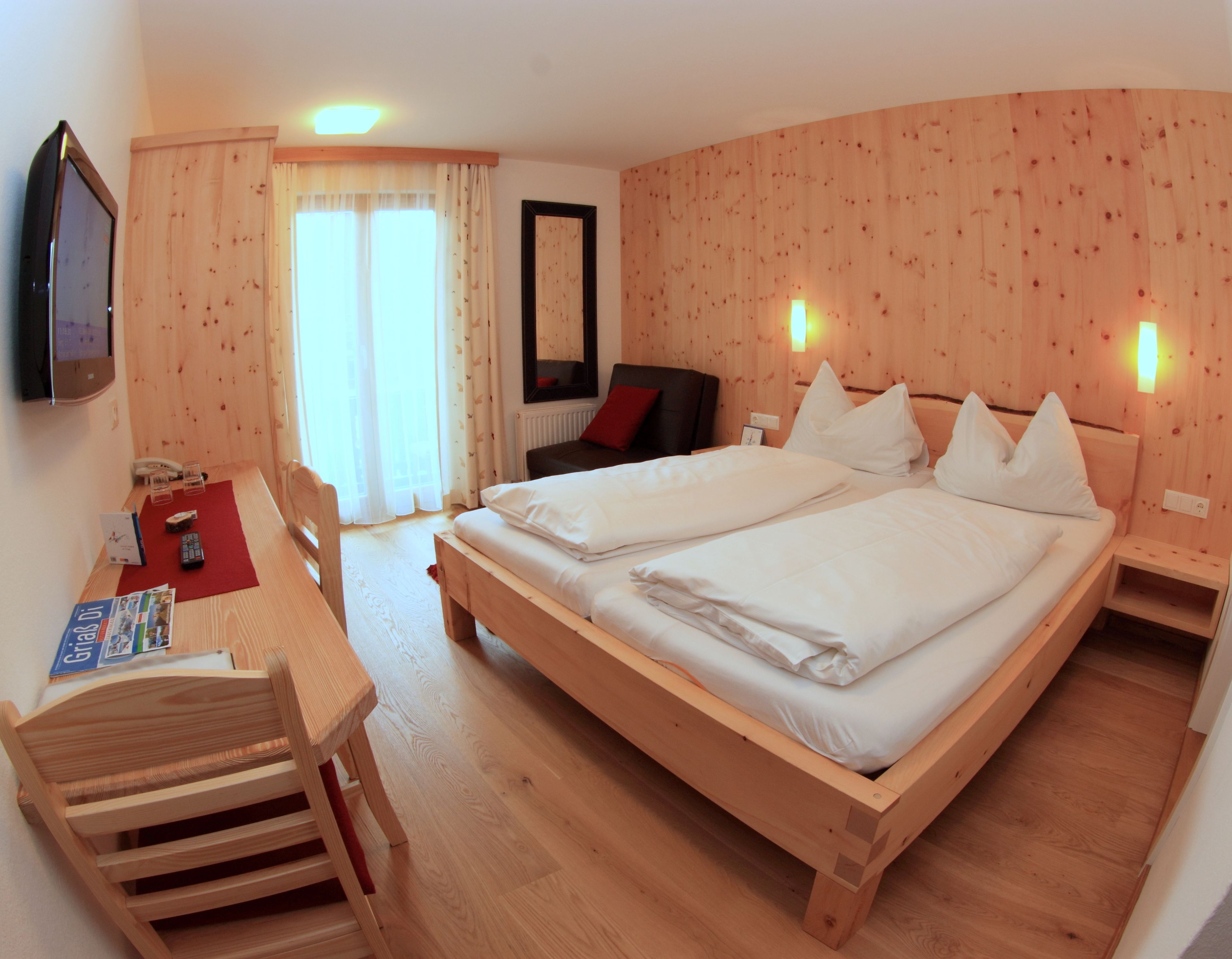 A double bed with a wooden frame, a desk with a chair, a television on the wall, and a window with curtains.