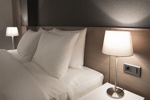 Een net opgemaakt bed met witte lakens en kussens, een bedlampje en een stopcontact.