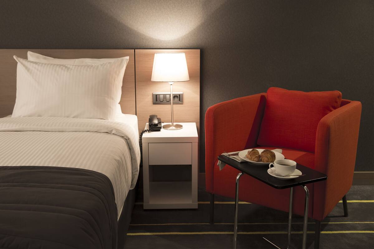 Une chambre d’hôtel avec un lit, une table de chevet avec une lampe, un fauteuil rouge et une petite table sur laquelle se trouvent une assiette de croissants et une tasse de café.
