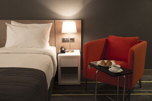 Hotelkamer met een bed, een nachtkastje met een lamp, een rode fauteuil en een kleine tafel met een bord croissants en een kop koffie.